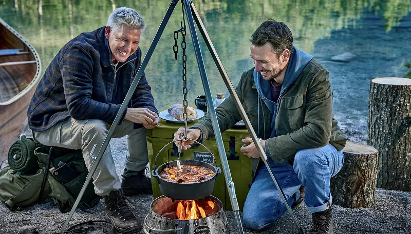 Bastian Schweinsteiger und Felix Neureuther kochen am See über einer Feuerschale einen Eintopf in einem gusseisernen Topf an einem Dreibein.